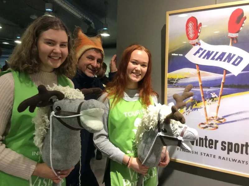 Kaksi naista ja kettupipoinen Timo keppihervosten kanssa suomi-julisteen edessä